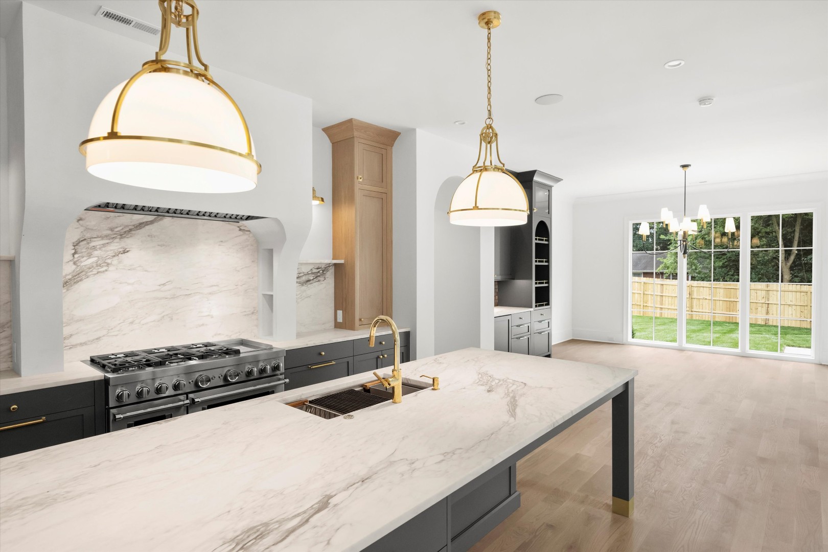 Custom Kitchen with Marble Island and Designer Lighting Bright custom kitchen featuring a marble waterfall island, brass pendant lighting, professional gas range, and open layout overlooking the backyard